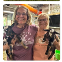 Mary and another smiling woman holding young goats in the barn.